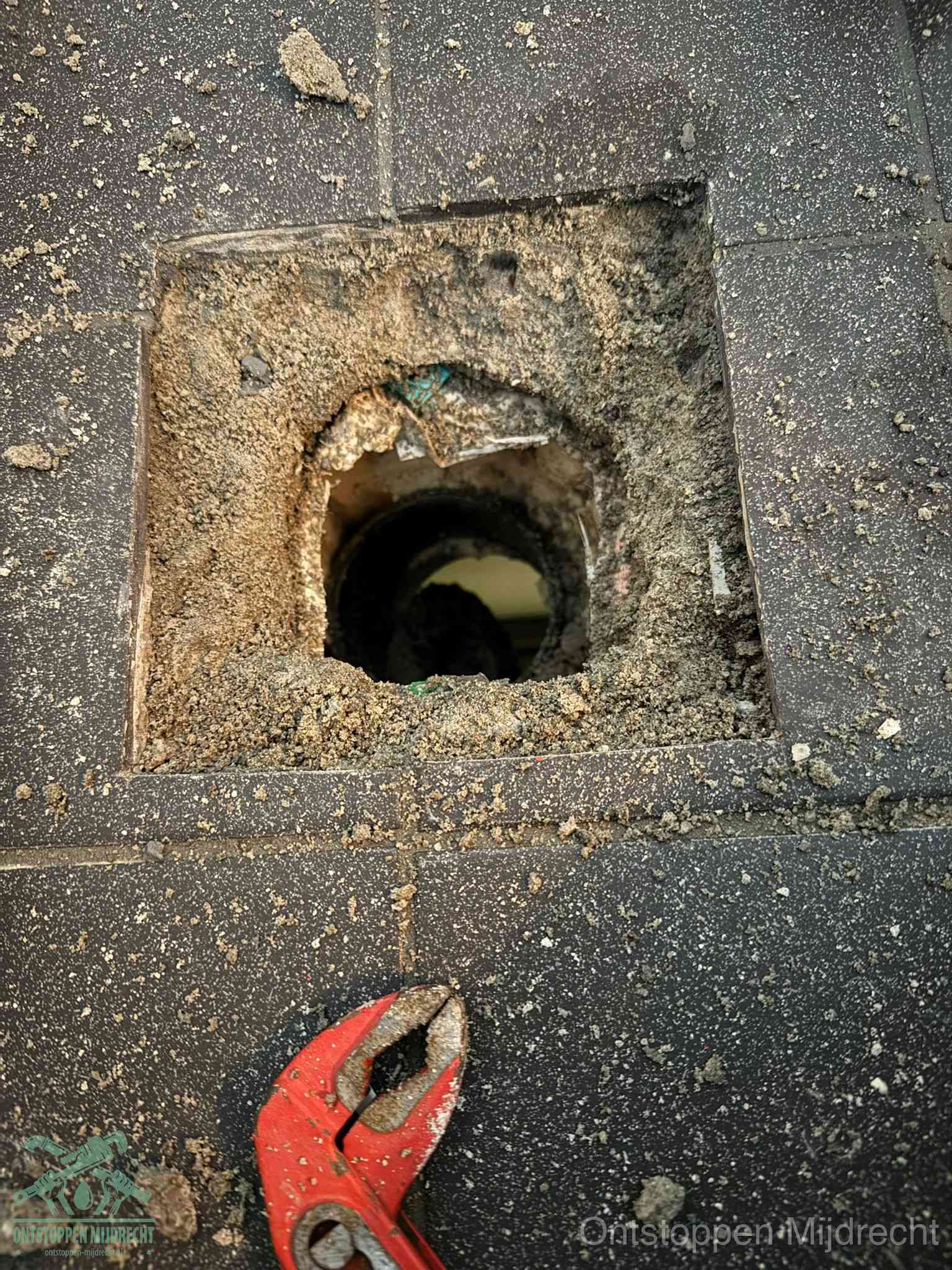 Een geopend rioolputje in een betontegelvloer met losgemaakt rooster en een zichtbare ronde afvoerbuis, het achterhalen waar de afvoerverstopping zit in Mijdrecht