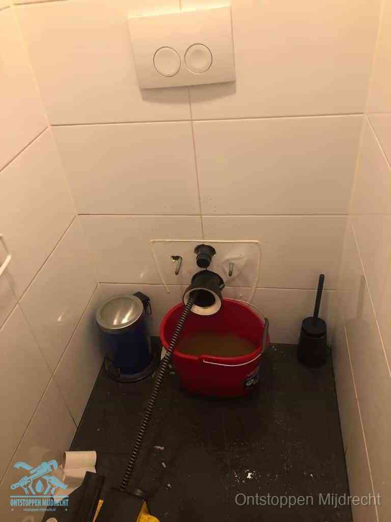 Een ontstopte wand-wc met verwijderde toiletpot waar een ontstoppingsveer in de wanduitgang verdwijnt, het achterhalen waar de verstopping zit in Mijdrecht