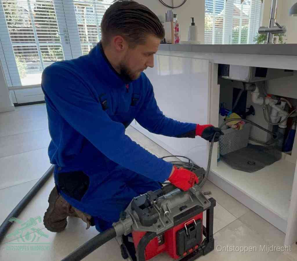 Een monteur met rode handschoenen knielt in een keuken en voert een elektrische ontstoppingsveer in tijdens het professioneel afvoer ontstoppen in Mijdrecht